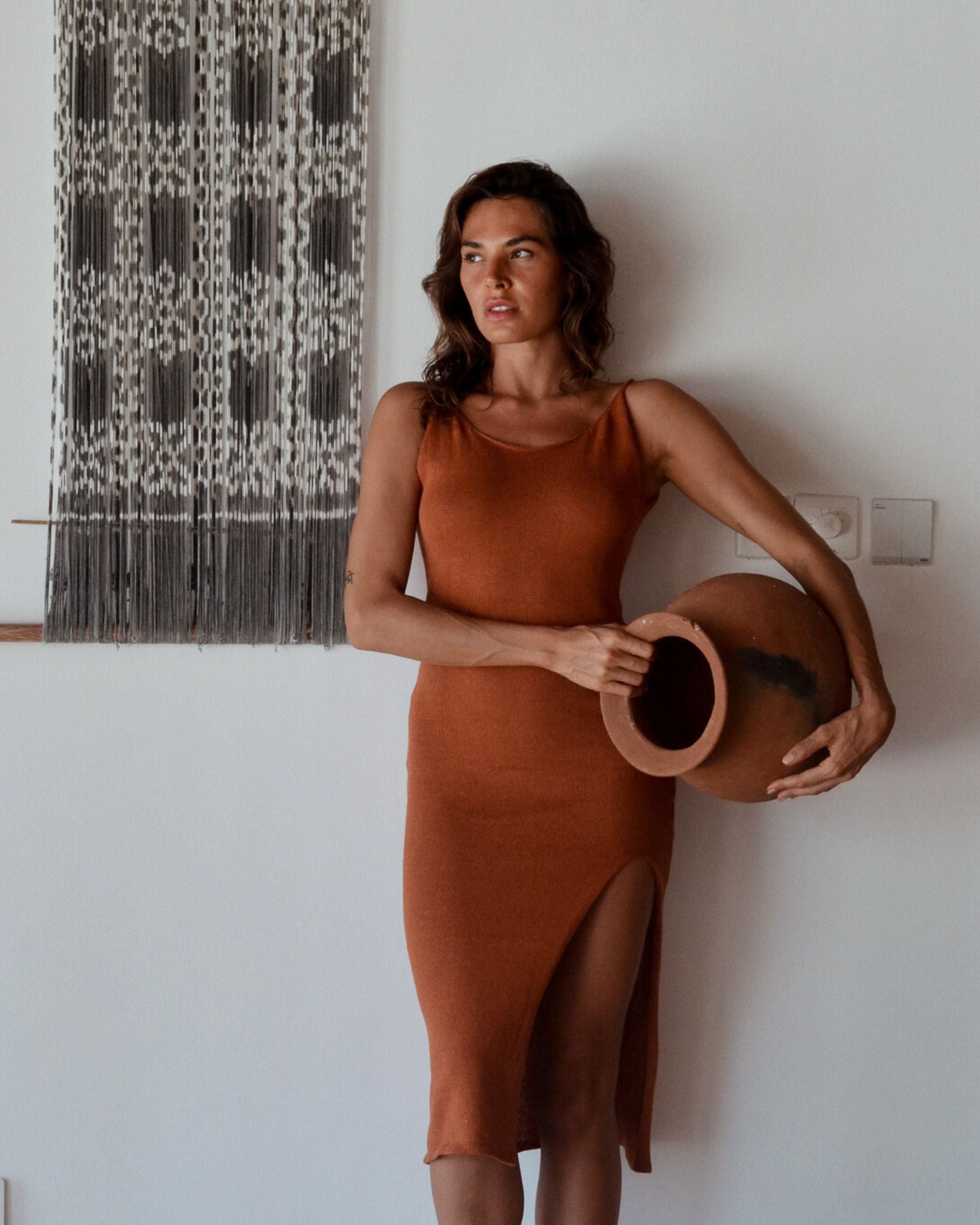 Woman in a rust-colored dress holding a ceramic pot against a white wall.
