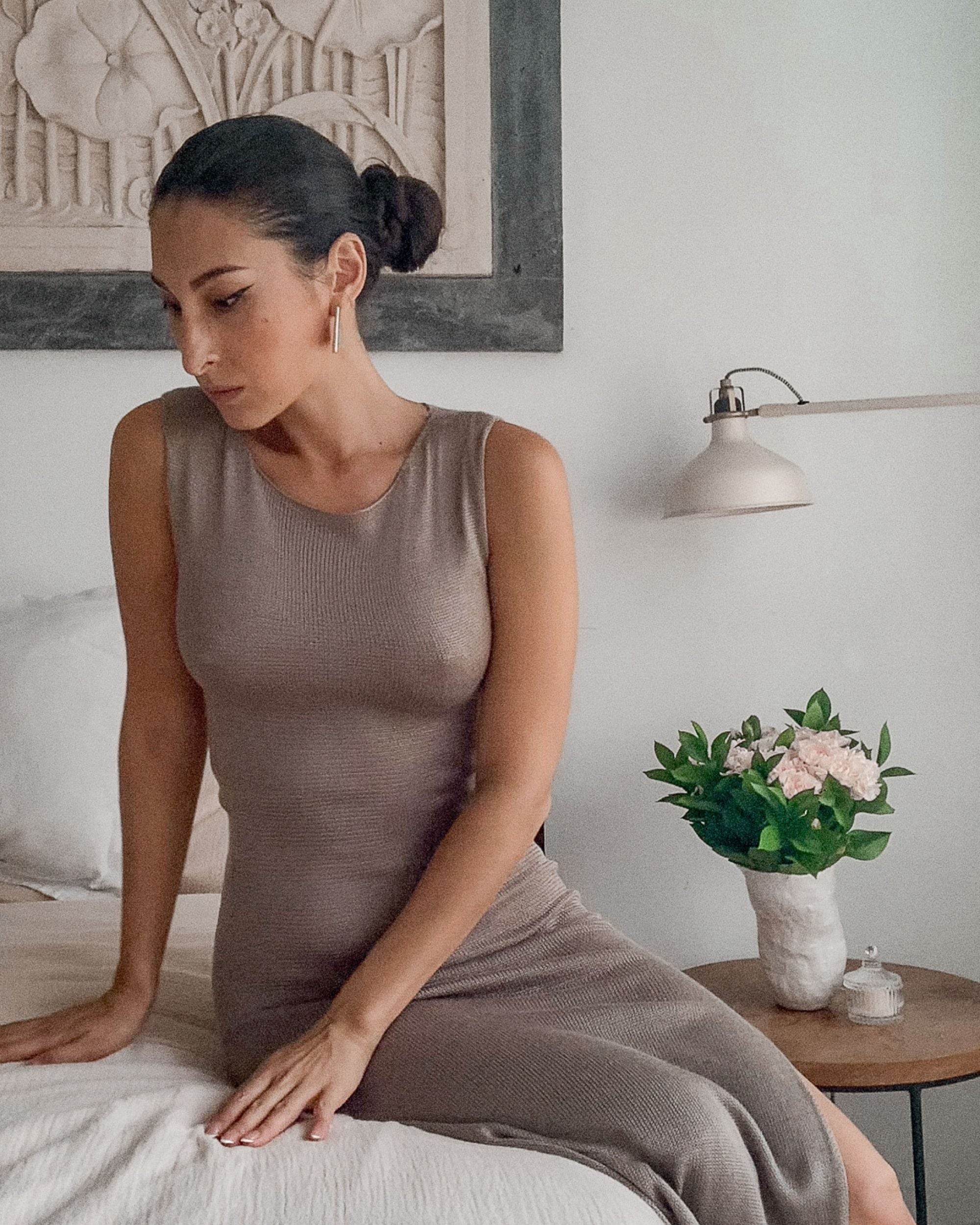 Woman in a brown dress sitting on a bed in a bedroom setting.
