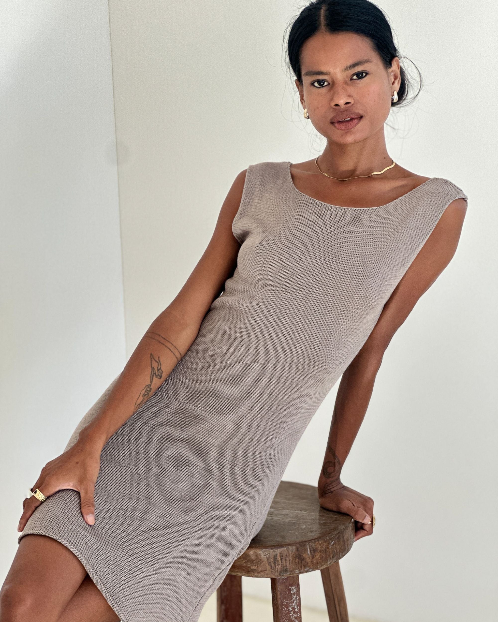 Woman wearing a beige sleeveless dress sitting on a wooden stool against a white background