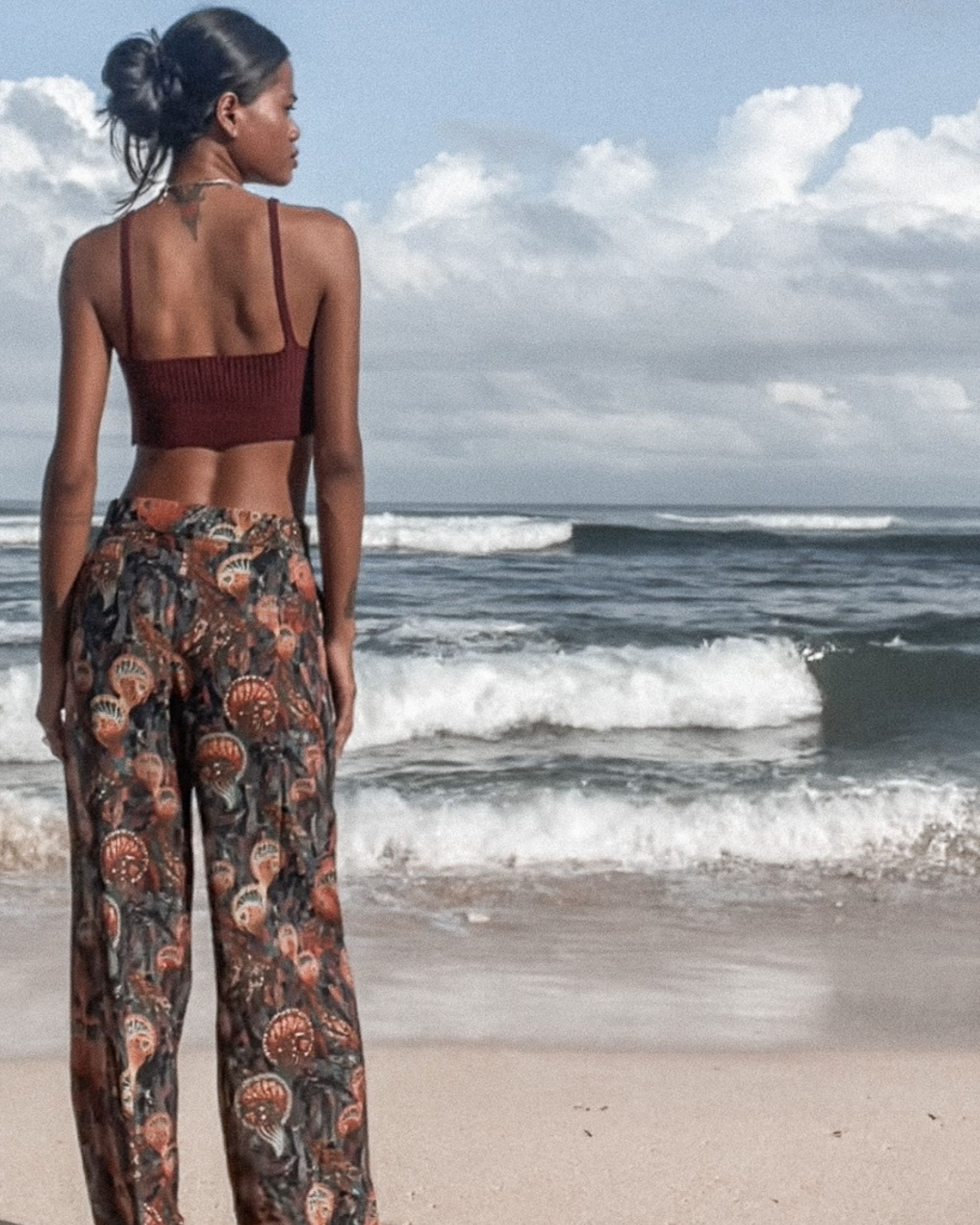 Woman standing on a beach wearing a patterned outfit, looking out at the ocean.